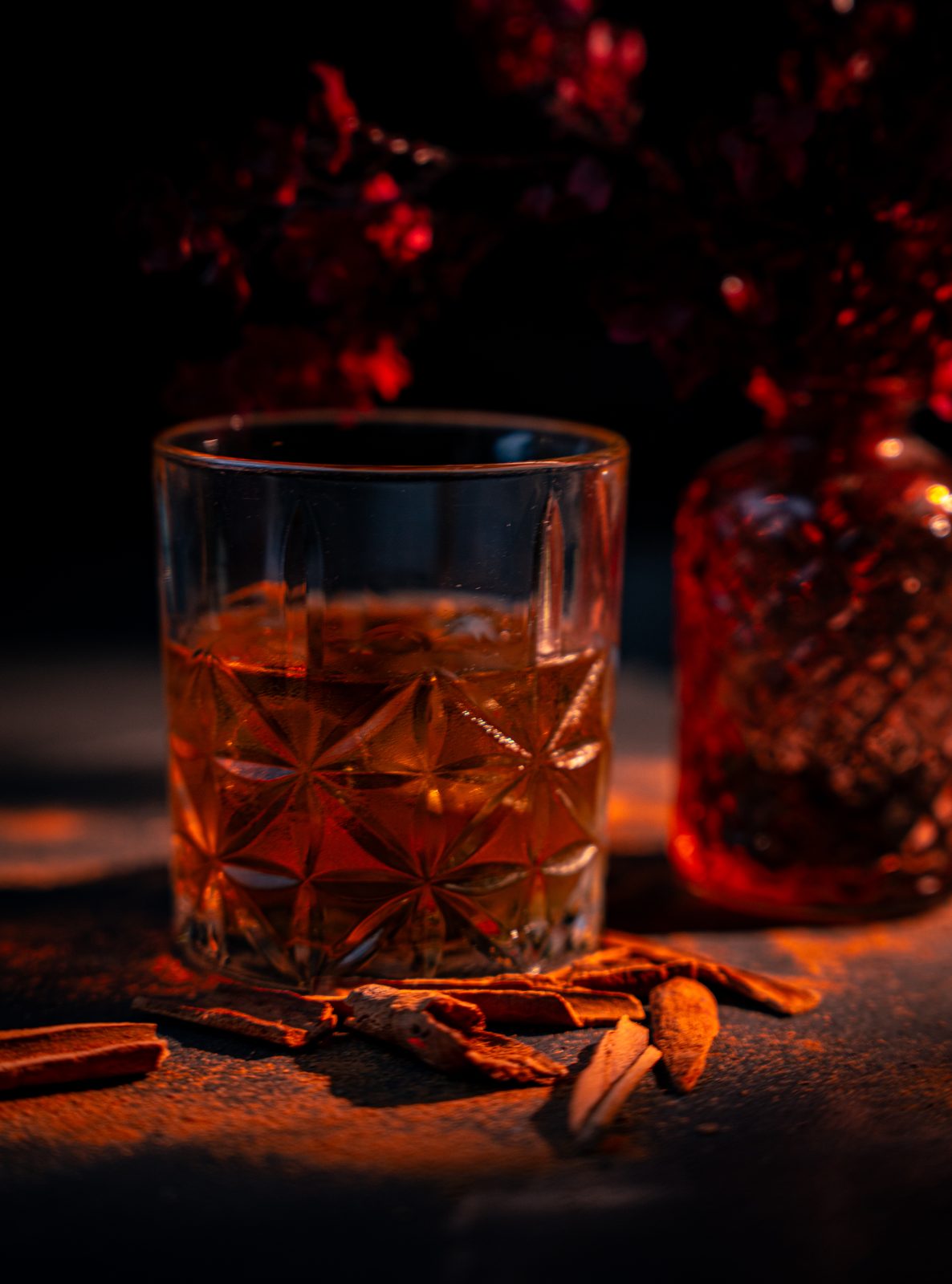 Ein Kristallglas mit bernsteinfarbenem Drink auf Eis, daneben Zimtstangen, im warmen gedämpften Licht