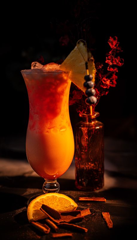 Cocktail im Hurricane-Glas mit Farbverlauf von Rot zu Orange, Ananasscheibe und Beerenspieß